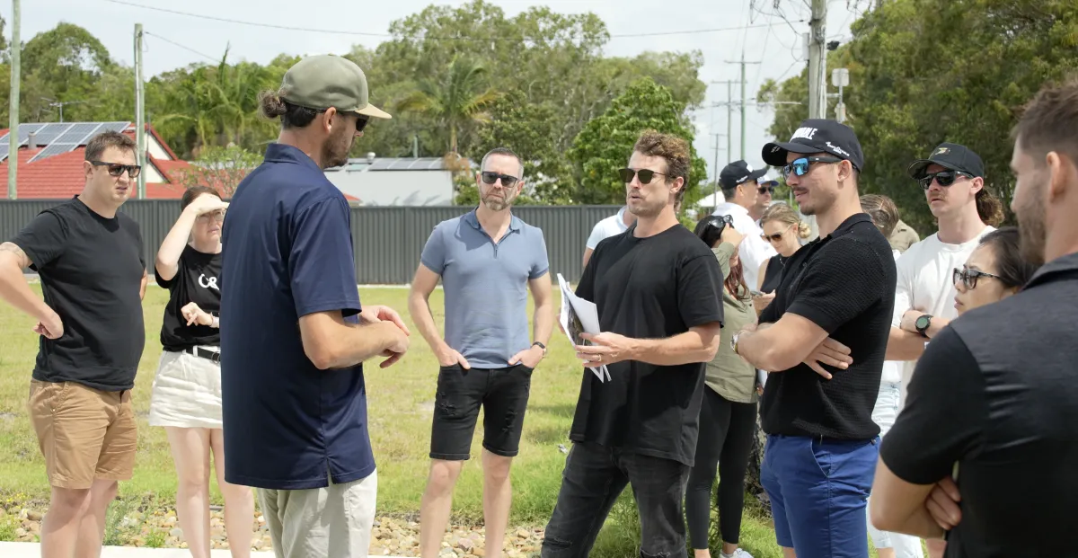A coach in a black t-shirt holding printed plans, talking through a site with a dozen students standing on a grassed block